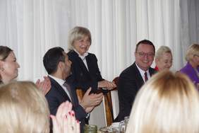 Pokaż zdjęcie: Zbliżenie na uczestników siedzących przy stole podczas oficjalnej części spotkania. W centrum starsza kobieta w czarnym żakiecie i białej bluzce z koronkowym kołnierzem uśmiecha się, patrząc w stronę pozostałych gości. Obok niej siedzi mężczyzna w okularach i garniturze, który szeroko się uśmiecha. Na pierwszym planie widoczne są rozmyte sylwetki innych osób klaszczących dłońmi, co sugeruje radosną i podniosłą atmosferę wydarzenia.