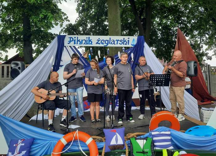 Pokaż zdjęcie: Prowizoryczna scena na podwyższeniu, na której stoi 6 osób w trakcie występu artystycznego. W tle na niebiesko-białej szarfie napis "Piknik żeglarski" Całość odbywa się w ogrodzie wśród drzew.