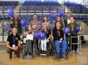 Pokaż zdjęcie: na zdjęciu hala sportowa, zwycięzcy turniejy pozują do zdjęcia trzymając w ręku dyplomy oraz balony z logo Fundacji Aktywnej Rehabilitacji. Część osób siedzi na wózkach inwalidzkich   