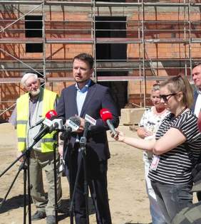 Fotografia plenerowa będąca zbliżeniem na centralną postać mężczyzny w garniturze przemawiającego podczas konferencji prasowej. Mężczyzna ma dłonie splecione przed sobą i patrzy w stronę mikrofonów. Po jego lewej stronie częściowo widoczny mężczyzna w odblaskowej kamizelce, a po prawej kobieta trzymająca kask i kolejny mężczyzna. Na pierwszym planie widać kilka mikrofonów różnych stacji telewizyjnych na statywach i ramię osoby trzymającej kostkę mikrofonową. W tle widać fragmenty rusztowań i ceglanego budynku w budowie pod błękitnym niebem.