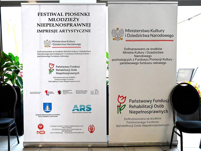 Stoją dwa banery: ten po lewo zawiera nazwę imprezy, jej organizatora - Fundację Omnibus - oraz sponsorów - ich nazwy i loga, w tym PFRON-. Na banerze po prawoa na tym na prawo informuje, że Festiwal został dofinansowany ze środków Ministerstwa Kultury i Dziedzictwa Narodowego oraz Państwowego Funduszu Rehabilitacji Osób Niepełnosprawnych