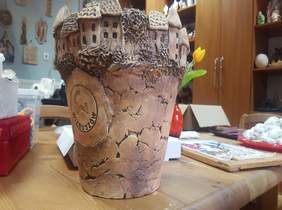 Pokaż zdjęcie: na zdjęciu duża gliniana donica wykończona w kształcie domków z herbem Wieruszowa w kolorze naturalnym glinianym