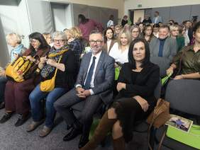 Pokaż zdjęcie: Zbliżenie na osoby siedzące w pierwszym rzędzie, w obiektyw patrzą mężczyzna w garniturze oraz młoda kobieta.