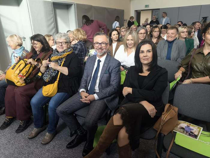 Pokaż zdjęcie: Zbliżenie na osoby siedzące w pierwszym rzędzie, w obiektyw patrzą mężczyzna w garniturze oraz młoda kobieta.