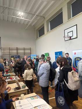 Pokaż zdjęcie: Grupa ludzi stoi przy stoiskach informacyjnych w sali gimnastycznej z koszem do koszykówki na ścianie; uczestnicy rozmawiają i przeglądają materiały na stołach.