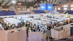 Pokaż zdjęcie: Szerokie ujęcie z góry na halę widowiskowo-sportową Enea Arena podczas targów pracy, wypełnioną licznymi białymi stoiskami wystawienniczymi i tłumem odwiedzających.