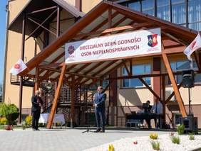 Przed budynkiem drewniana konstrukcja w formie dachu, duży napis diecezjalny dzień  godności osób niepełnosprawnych, mężczyzna przed mikrofonem przemawia, z lewej strony drugi mężczyzna trzymający teczkę, skierowany w stronę przemawiającego, słoneczny dzień