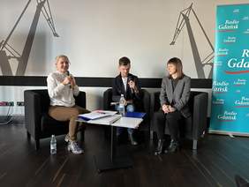 Pokaż zdjęcie: Trzy dwie kobiety i mężczyzna siedzą na fotelach. Kobieta po lewej stronie w blond włosach trzyma mikrofon i mówi. Za nimi rysunki dźwigów stoczni gdańskiej. Po prawej baner Radia Gdańsk.