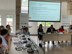 Pokaż zdjęcie: Jasna sala konferencyjna z dużymi oknami. Stoły konferencyjne z jasnego drewna, na których leżą mikrofony i dokumenty. Za nimi siedzą ludzie, rozmawiają. W prawym rogu telebim  na którym widnieje slajd z tytułem „Samodzielność-Aktywność-Mob