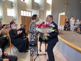 Pokaż zdjęcie: Ubrany w granatowy garnitur, biała koszule i krawat mężczyzna wręcza kobiecie ubranej w długą sukienkę kwiaty. W tle widownia i artyści bijący brawo.