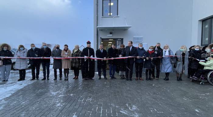 Pokaż zdjęcie: Obok siebie w jednej linii stoi kilkanaście osób trzymających biało czerwoną szarfę i nożyczki w ręku. Za nimi nowy budynek. Przed budynkiem duży plac z kostki brukowej.