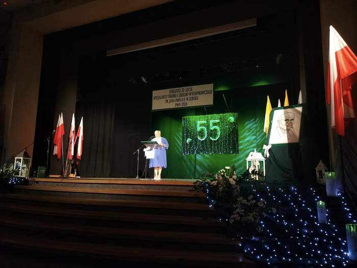 Pokaż zdjęcie: Na scenie, zaciemnionej, stoi Dyrektor SOSW - kobieta, ubrana na niebiesko, czyta przemówienie