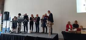 Pokaż zdjęcie: Sala konferencyjna, na scenie stoi grupa siedmiu osób, wysoki mężczyzna w garniturze wręcza grawerton kobiecie blondynce.