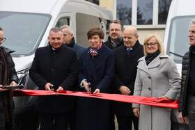 Na zdjęciu jest grupa osób podczas uroczystego przecięcia czerwonej wstęgi. Na fotografii widać kilka osób w płaszczach stojących w rzędzie; dwie osoby trzymają nożyczki nad wstęgą. W tle stoją nowe busy dla Warsztatów Terapii Zajęciowej w Grodzisku Wielkopolskim i Rakoniewicach