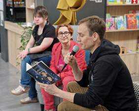 Pokaż zdjęcie: Aktor Mateusz Janicki czyta książkę "Konik i Koninka". Siedzi na fotelu. Obok niego siedzi kobieta z dorosłą córką z zespołem Downa.