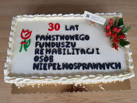 Prostokątny, jasny tort, przyozdobiony z lewej strony logiem PFRON, z prawej kwiatami u góry napis 30 lat, centralną cześć wypełnia napie, pełna nazwa PFRON