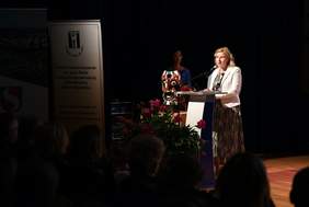 Pokaż zdjęcie: Na scenie przy mównicy stoi kobieta blondynka ubrana w czarno białą sukienkę oraz jasny żakiet. Przed mównicą stoi wazon z piwoniami