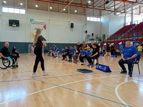 Pokaż zdjęcie: W hali sportowej trwa Turniej Boccia. Zawodnicy siedzą na krzesłach, a przed nimi leżą niebieskie i czerwone kule. 
