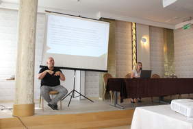 Pokaż zdjęcie: W sali konferencyjnym po lewej stronie siedzi na krześle tłumacz języka migowego, po prawej za stołem prezydialnym siedzi kobieta, druga stoi z mikrofonem, w tle po lewej na ekranie wyświetlana jest prezentacja