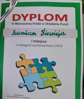 Pokaż zdjęcie: Kartka papieru z zielonym obramowaniem zawierająca następujące sformułowania: „Dyplom”, „Mistrzostwa Polski w Układaniu Puzzli”, „Damian Siewieja”, „I miejsce w kategorii wychowanków ZPEW” oraz „Organizatorzy: AS, Leśny Zakątek, Foto Olszewscy”. Na dyplomie widoczne są również połączone ze sobą cztery puzzle.