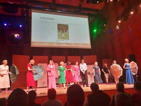 Pokaż zdjęcie: Na scenie stoi sześć osób z mikrofonami w rękach. Z prawej i lewej strony stoją po dwie panie z kolorowymi parasolkami w rękach. Nad nimi zawieszony jest ekran, na którym wyświetlony jest obraz. 