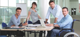 Pokaż zdjęcie: Wokół biurka znajdują się cztery osoby: jedna kobieta i trzech mężczyzn. Jeden z panów siedzi na wózku inwalidzkim. Wszyscy patrzą w stronę obiektywu aparatu.  Na biurku leżą dokumenty, telefon, laptop. 