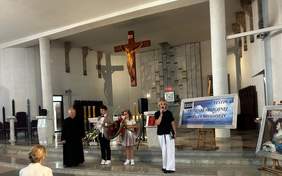 Pokaż zdjęcie: Wnętrze kościoła podczas festiwalu piosenki religijnej. Na ołtarzu stoi ksiądz, obok chłopiec i dziewczynka z kwiatami, oraz kobieta przemawiająca do mikrofonu. W tle widoczny jest marmurowy ołtarz oraz dwa plakaty informujące o XXIV Podlaskim Festiwalu Piosenki Religijnej Dzieci i Młodzieży z Niepełnosprawnością Intelektualną.