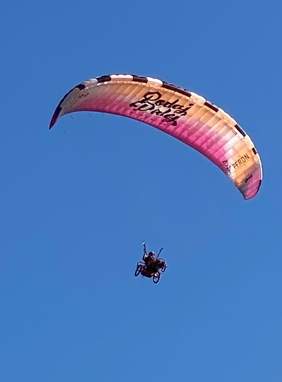 Pokaż zdjęcie: Uczestnik z przypiętym do wózka inwalidzkiego paralotnią leci na tle błękitnego niebie