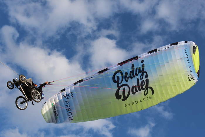Pokaż zdjęcie: Zdjęcie przedstawia osobę na wózku, która leci pod skrzydłem paralotniowym. Nad osobą rozciąga się duży, kolorowy żagiel paralotni z wyraźnym napisem „Fundacja Podaj Dalej”. Na paralotni można też dostrzec logo „PFRON”. W tle widoczne jest niebo z białymi chmurami. Osoba na wózku trzyma linki sterujące paralotnią