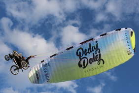 Pokaż zdjęcie: Zdjęcie przedstawia osobę na wózku, która leci pod skrzydłem paralotniowym. Nad osobą rozciąga się duży, kolorowy żagiel paralotni z wyraźnym napisem „Fundacja Podaj Dalej”. Na paralotni można też dostrzec logo „PFRON”. W tle widoczne jest niebo z białymi chmurami. Osoba na wózku trzyma linki sterujące paralotnią