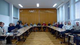 Pokaż zdjęcie: Zdjęcie przedstawia dużą, jasną salę konferencyjną z długim stołem ustawionym w literę „U”. Przy stole siedzi kilkanaście osób, zarówno kobiety, jak i mężczyźni, w różnym wieku. Wszyscy są zwróceni w stronę fotografa, patrzą w obiektyw i niektórzy się uśmiechają. Na stole znajdują się laptopy, notatki, butelki z wodą oraz szklanki. W tle widać zasłonięte pomarańczowe zasłony oraz duże okna z białymi roletami. Podłoga jest drewniana, a światło w pomieszczeniu jest jasne.