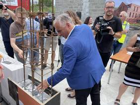 Pokaż zdjęcie: Mężczyzna w niebieskiej marynarce, ciemnych spodniami białej koszuli wrzuca biały kamyczek pomiędzy zbrojeniami do filaru.