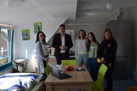 Pokaż zdjęcie: Mężczyzna i cztery kobiety znajdują się w mieszkaniu. Przed nimi stoi stół, a za nimi jest kuchnia