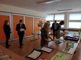 Pokaż zdjęcie: 5 osób 3 kobiety i dwóch mężczyzn ogląda prace plastyczne rozłożone na stołach