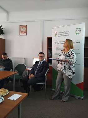 Pokaż zdjęcie: Trzy osoby. Dwie z nich siedzą na krzesłach, jedna na stojąco przemawia