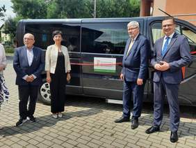 Pokaż zdjęcie: Przed samochodem przystosowanym do przewozu osób z niepełnosprwnością stoi kobieta i trzech mężczyzn. Na pojeździe naklejona jest naklejka z informacją, że auto zostało dofinansowane ze środków PFRON. 