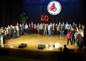 Pokaż zdjęcie: Na scenie stoją w półkolu ludzie. Wszyscy mają przewieszone przez szyje medale. W tle wisi dekoracja z napisem: Warsztat Terapii Zajęciowej w Drzewicy. 