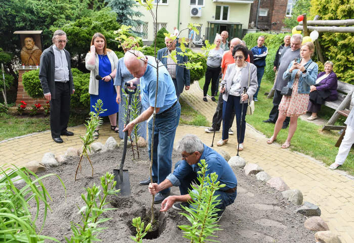Pokaż zdjęcie: Zdjęcie grupy osób w parku, dwie z nich sadzą dąb, wokół stoi grupa zaproszonych gości i się przygląda pracom