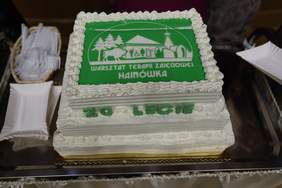 Pokaż zdjęcie: Prostokątny tort z białym kremem i zieloną dekoracją wierzchnią z logo Warsztatu Terapii Zajęciowej w Hajnówce oraz napisem „20-lecie”.