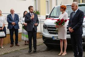 Pokaż zdjęcie: Dyrektor Oddziału trzyma w ręku mikron, stojąc na tle nowe auta, obok są inne osoby, i mówi w stronę Minister Elżbiety Rafalskeij