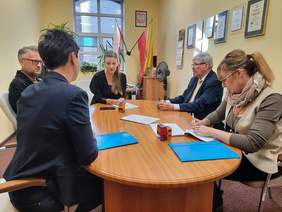 Pokaż zdjęcie: Przy owalnym stole siedzi pięć osób: trzy kobiety i dwóch mężczyzn. Rozmawiają ze sobą i podpisują dokumenty. 