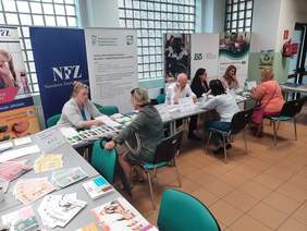 Pokaż zdjęcie: Przy trzech różnych stoiskach informacyjnych siedzą eksperci, którzy udzielają informacji osobom zainteresowanym. Za nimi stoją banery reklamowe różnych instytucji. 