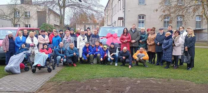 Pokaż zdjęcie: Mieszkańcy DPS, wśród których są osoby na wózku, stoją na tle nowego auta, są ubrani na zimowo i są uśmiechnięci i weseli