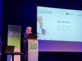 Pokaż zdjęcie: Prelegentka stoi przy mównicy ZUS na oświetlonej scenie, za nią ekran z prezentacją „Dni Seniora 2025” i grafiką dotyczącą bezpieczeństwa seniorów.