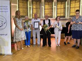 Pokaż zdjęcie: Na scenie stoi dwóch mężczyzn i pięć kobiet. Jeden z nich ubrany w jasne spodnie i koszulę trzyma dyplom oprawiony w ramę. Drugi obok w okularach białej koszuli i kamizelce trzyma kwiaty. Po bokach dwie kobiety również trzymają ramkę i kwiaty. 