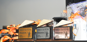 Pokaż zdjęcie: Na stole ustawione są trzy statuetki dla laureatów I edycji Warsztat Film Festiwalu z wygrawerowaną nazwą festiwalu, zajętym miejscem, kamerą oraz napisem: „Leszno 05.06.2024”. Obok statuetek widoczne są także nagrody rzeczowe.