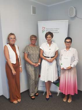 Pokaż zdjęcie: W rogu sali szkoleniowej stoją cztery kobiety na tle baneru PFRON. Powyżej baneru znajduje się kratka wentylacyjna oraz głośnik. Ściany sali są w kolorze białym.