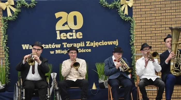 Pokaż zdjęcie: Grupa pięciu mężczyzn siedzi na scenie i gra na trąbkach. W tle, na granatowym płótnie udekorowanym dwoma złotymi kokardami oraz zielonymi łańcuchami widnieje napis: „20-lecie Warsztatów Terapii Zajęciowej w Miejskiej Górce”.