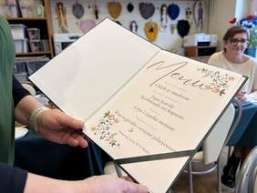 Pokaż zdjęcie: Menu w eleganckiej oprawie, trzymane przez osobę w zielonym stroju. W menu znajdują się dania, takie jak chleb ze smalcem, zupa śląska, karbinada bez kapusty oraz deser z ciastek owsianych. W tle widać osobę przy stole.