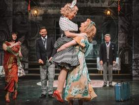 Pokaż zdjęcie: Scena teatru podczas przedstawienia. Na pierwszym planie jedna aktorka podnosi drugą w górę, za nimi stoą dwaj mężczyźni, a obok mężczyzna przebrany za kobietę.